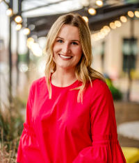 Headshot - Ali Jeffries White woman with blond hair in red top smiling in front of outdoor awaning with lights