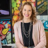 A white woman with dark blond hair, dressed in a black top and pink cardigan, sitting in front of a colorful wall at a desk with magazines and an open laptop.