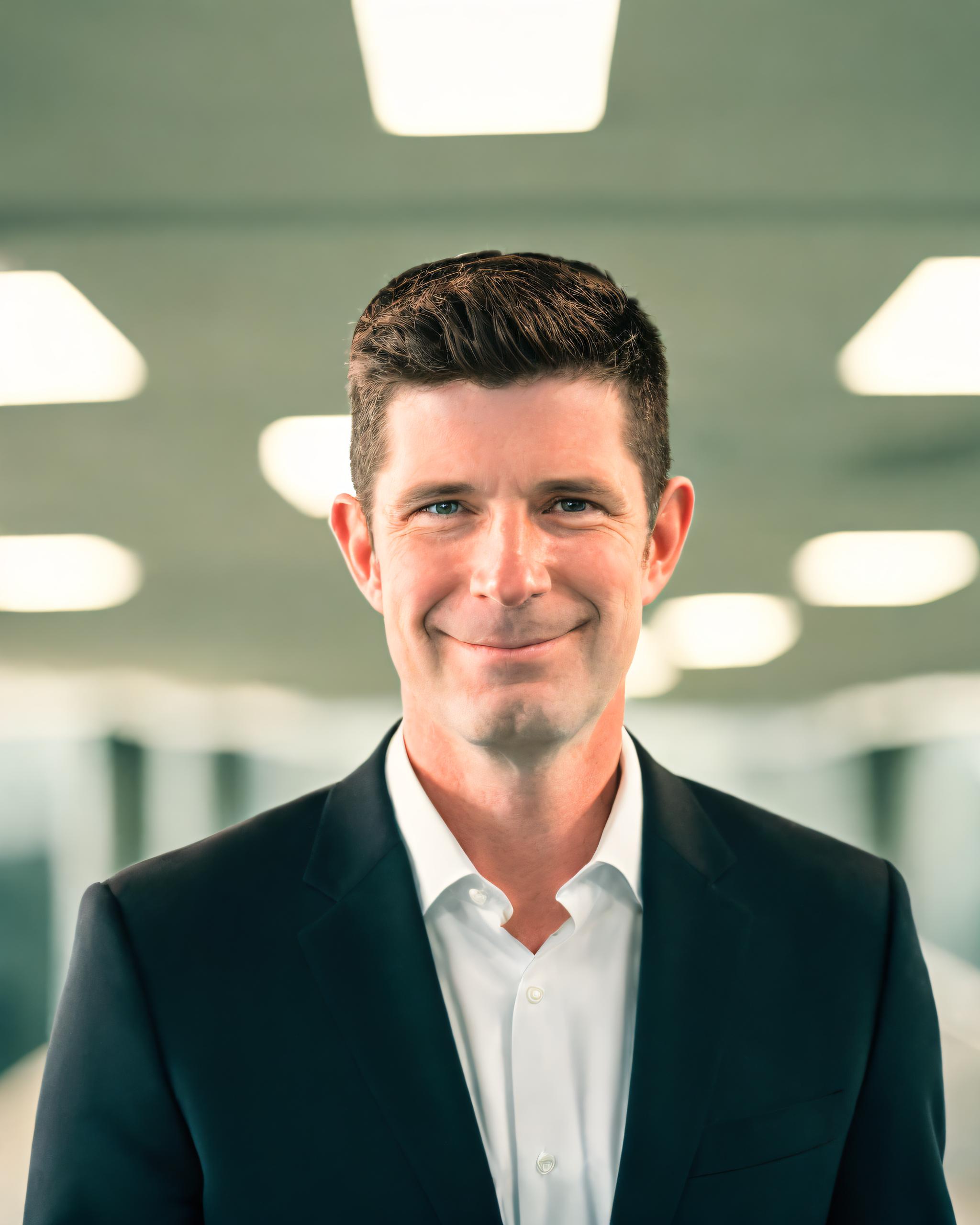 White man with goatee in black blazer and white collared shirt standing in an office with overhead flourescent lights